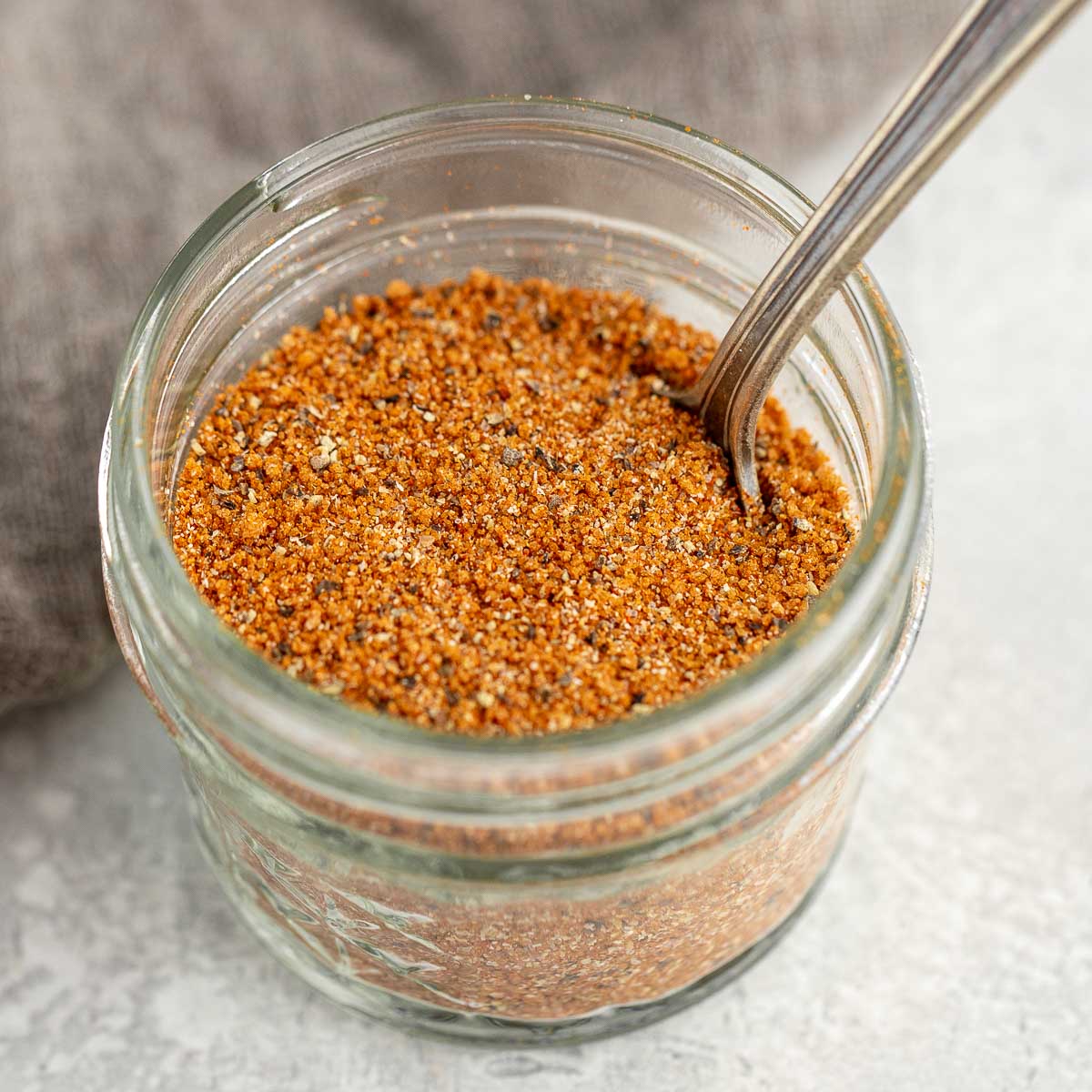 A small glass jar holding the finished bbq rub with a spoon to scoop some out.