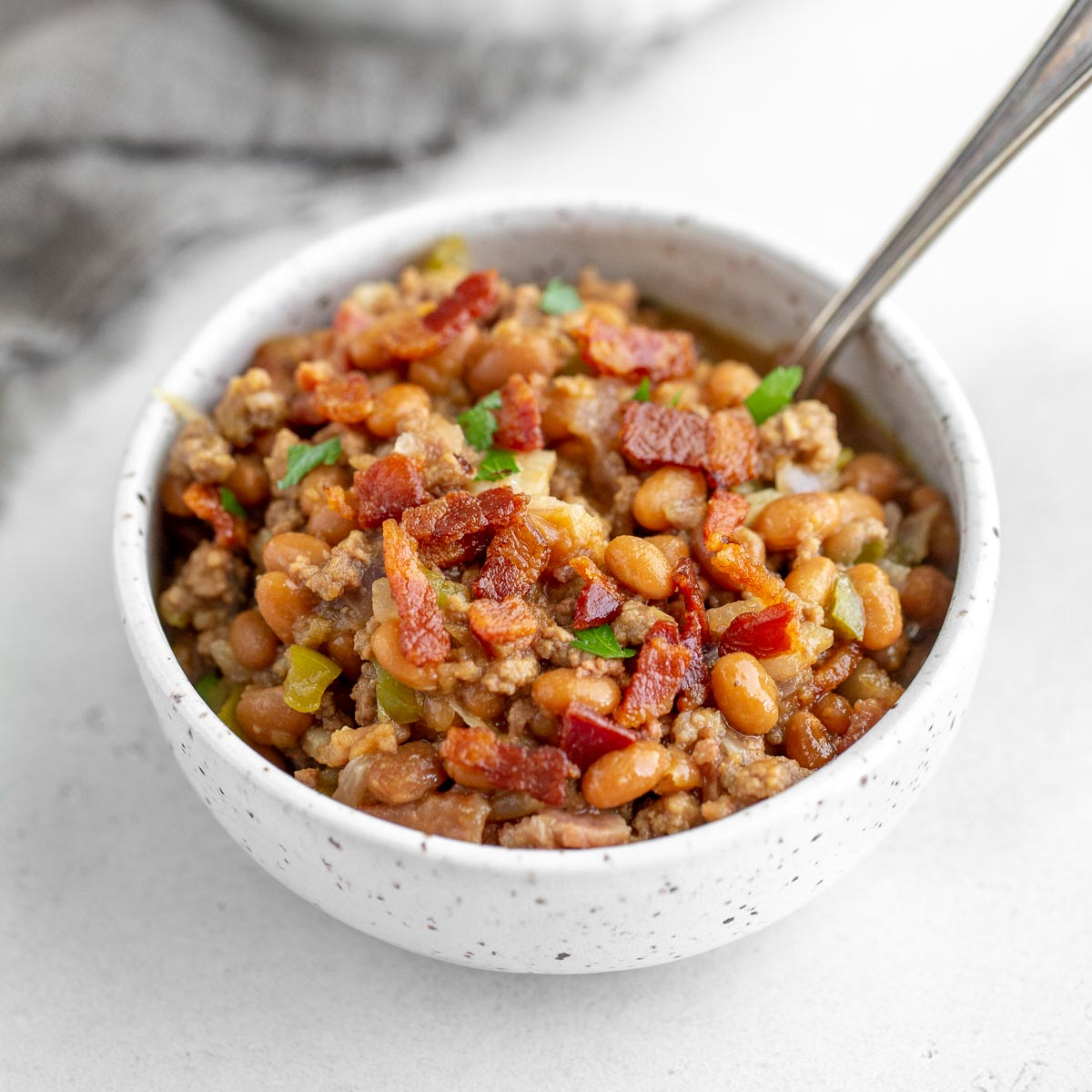 A speckled white bowl filled with baked beans and topped with bacon and parsley.