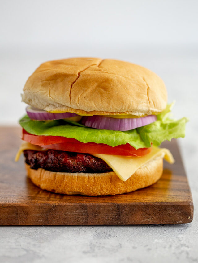 A fully assembled hamburger with cheese and vegetables sitting on a wood tray.