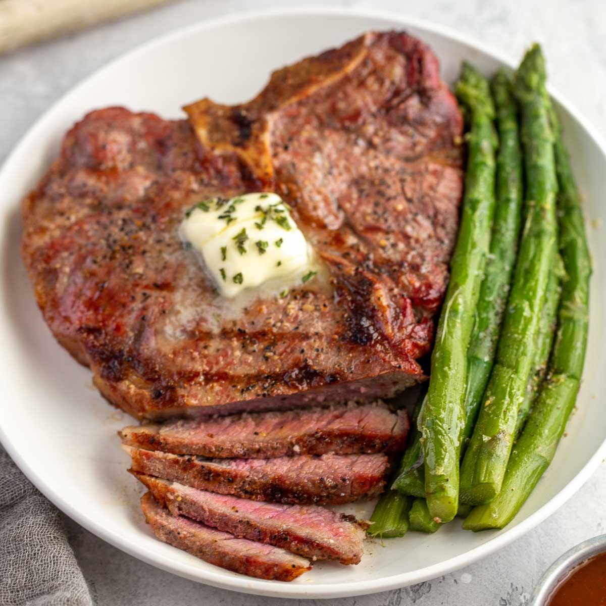 A grilled T-bone steak topped with herb butter on a white plate with a side of asparagus.