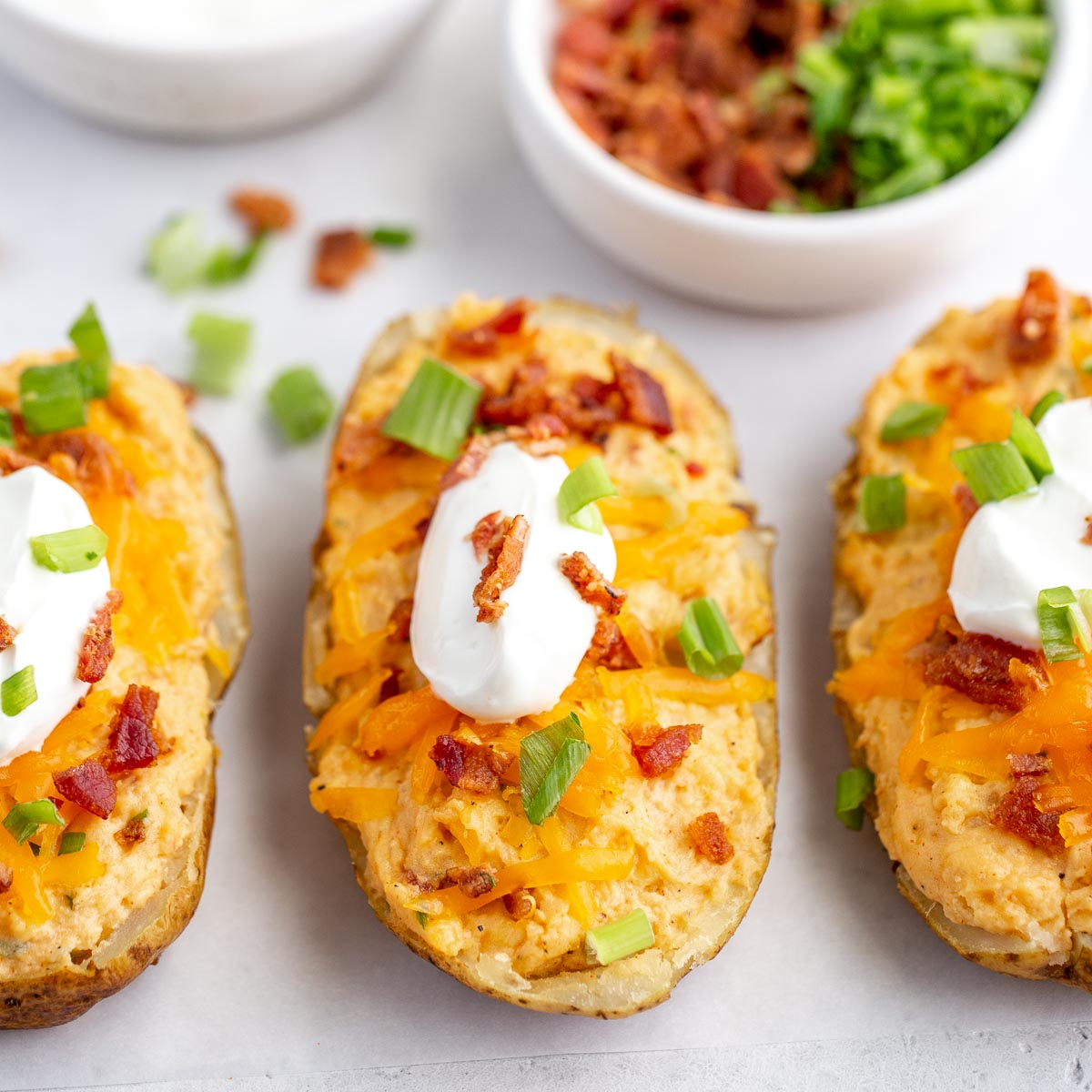 Twice baked potatoes topped with sour cream, bacon, and green onions on a gray surface with more sour cream and toppings in the background.